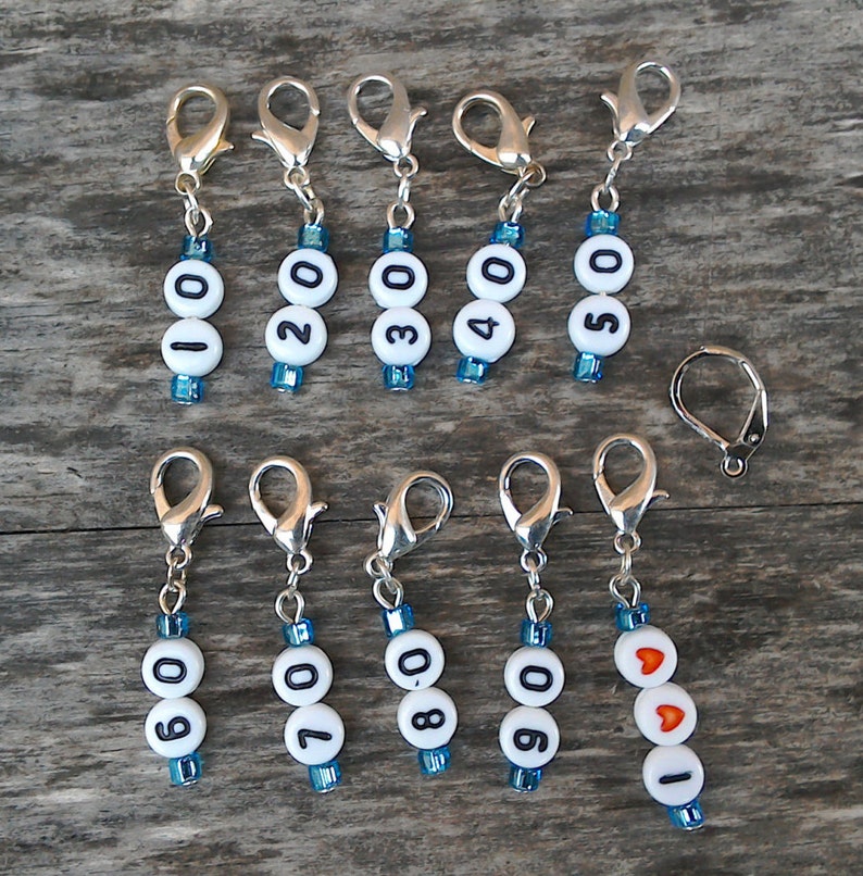May include: Ten silver stitch markers with white beads and black numbers 0-9 and one silver stitch marker with white beads and a red heart. The stitch markers are on a wooden surface.