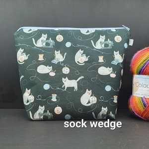 Può includere: Una pochette verde con cerniera con un motivo ripetuto di gatti che cuciono e tengono gomitoli di lana. La pochette è etichettata "sock wedge".