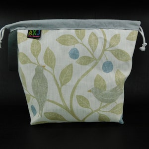 May include: A white drawstring bag with a blue and green floral pattern featuring birds and berries. The bag has a black drawstring and a label that reads "AXJ amelia.x.joy".
