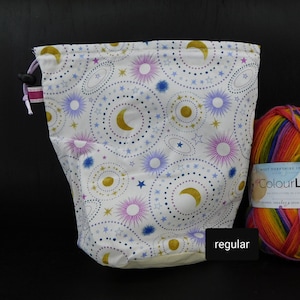 May include: White drawstring project bag with a moon and stars pattern. The bag is made of fabric and has a drawstring closure. The bag is sitting on a table next to a ball of rainbow yarn.