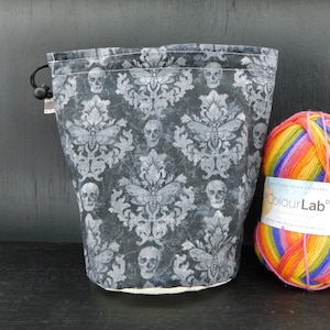 May include: A black drawstring project bag with a skull and moth pattern. The bag is sitting next to a ball of rainbow yarn.