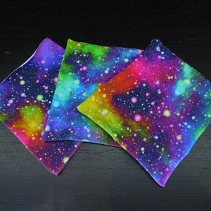 May include: Three square fabric coasters with a galaxy print. The coasters feature a dark blue background with bright pink, green, and yellow nebulae and white stars.