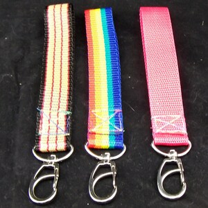 Puede incluir: Tres llaveros con correas rayadas de diferentes colores y clips giratorios plateados. Las correas son roja y blanca, arcoíris y rosa.