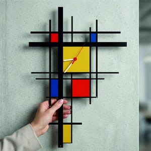 Puede incluir: Un reloj de pared con una cuadrícula geométrica de líneas negras finas que se cruzan con bloques rectangulares en colores primarios rojo, amarillo y azul. La esfera incluye manecillas blancas y un segundero fino de color rojo.