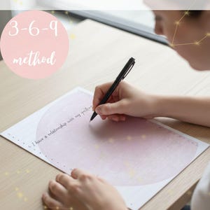 Può includere: Una pagina di manifestazione con il testo "Manifestation Page" in blu. La pagina ha un cerchio rosa con "3-6-9 method" scritto in bianco. Una persona sta scrivendo sulla pagina con una penna nera. La pagina ha un design rosa chiaro.