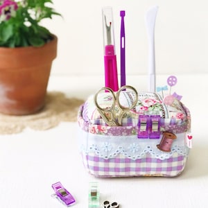 May include: A purple and white gingham fabric box with a floral design and lace trim. The box contains sewing supplies, including scissors, thread, pins, and a seam ripper. There are also several purple and green sewing clips and wooden spools of thread on a white surface.