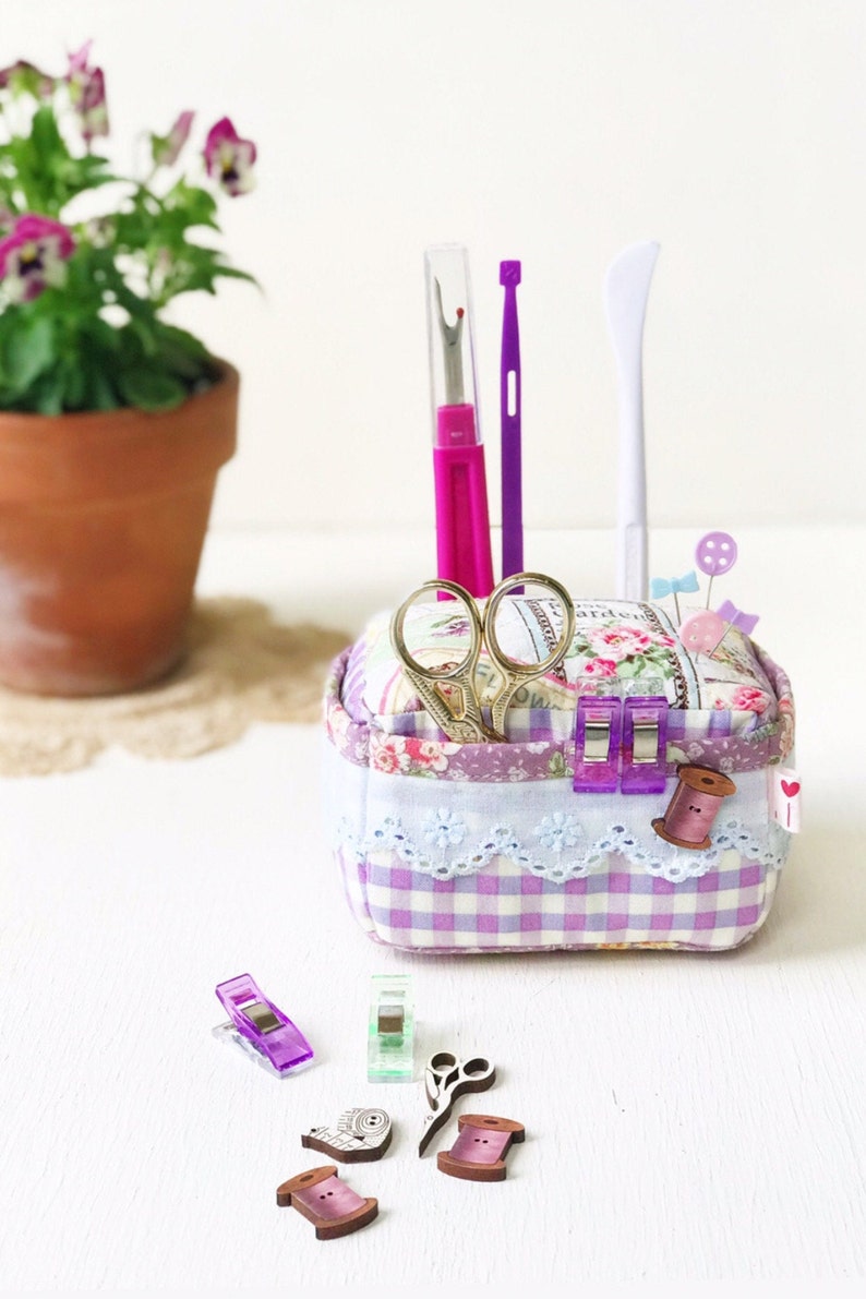 May include: A purple and white gingham fabric box with a floral design and lace trim. The box contains sewing supplies, including scissors, thread, pins, and a seam ripper. There are also several purple and green sewing clips and wooden spools of thread on a white surface.