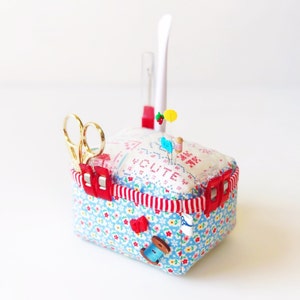 May include: A white and blue fabric pincushion with red trim and a white fabric top with the word "SUITE" embroidered in red. The pincushion has a red plastic clip on the side and a pair of gold scissors in the front. There are several colourful pins and a spool of thread on top of the pincushion.