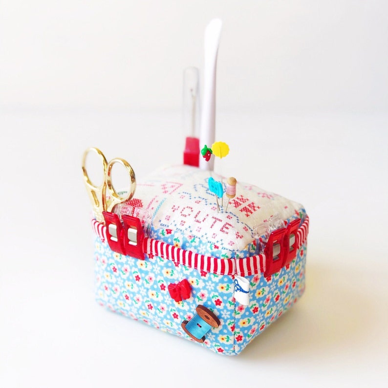 May include: A white and blue fabric pincushion with red trim and a white fabric top with the word "SUITE" embroidered in red. The pincushion has a red plastic clip on the side and a pair of gold scissors in the front. There are several colourful pins and a spool of thread on top of the pincushion.