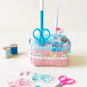May include: A pink fabric pincushion with a blue and white striped trim. The pincushion is filled with pins and needles. There are two pairs of scissors, a blue pen, and a white tool. The pincushion is sitting on a white surface with two spools of thread and three plastic bows.