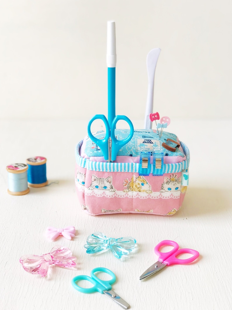 May include: A pink fabric pincushion with a blue and white striped trim. The pincushion is filled with pins and needles. There are two pairs of scissors, a blue pen, and a white tool. The pincushion is sitting on a white surface with two spools of thread and three plastic bows.