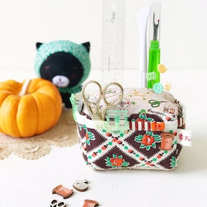 May include: A brown and white fabric sewing basket with a checkered pattern and red roses. The basket is filled with sewing supplies, including scissors, a needle threader, and buttons. There is a small pumpkin, a black and green stuffed animal, and a ruler in the background.