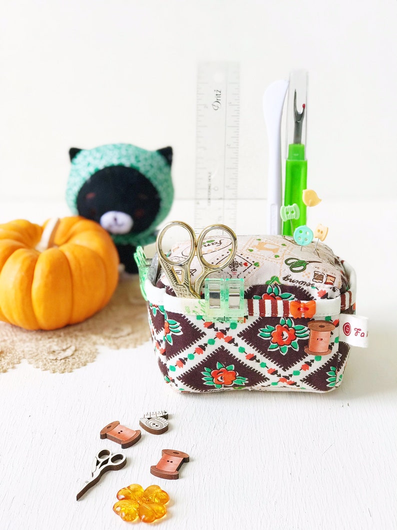May include: A brown and white fabric sewing basket with a checkered pattern and red roses. The basket is filled with sewing supplies, including scissors, a needle threader, and buttons. There is a small pumpkin, a black and green stuffed animal, and a ruler in the background.