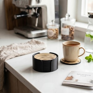 May include: A set of round coasters featuring a black holder and light wood inserts with a wavy, slatted design. The coasters are stacked inside the holder, with one individual coaster placed separately under a beige ceramic mug on a white surface.