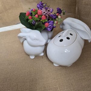 May include: Three white ceramic bunny-shaped vases. One vase holds a bouquet of purple, pink, and green artificial flowers. The other two vases are empty. The vases are on a brown burlap surface.