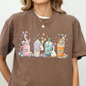 Può includere: T-shirt color espresso con una grafica colorata di bevande a tema pasquale. Il design include Hoppy Shake, Latte Joy, Boba Paradise e Carrot Craze, ognuno con un personaggio a forma di coniglietto. La maglietta è in stile casual e confortevole.
