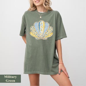 Puede incluir: Camiseta verde militar con un diseño de concha. La concha está compuesta por telas estampadas azules, amarillas y blancas. La camiseta tiene mangas cortas y un corte holgado.