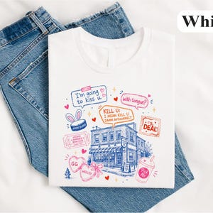 May include: A white t-shirt featuring a blue line-art illustration of a building surrounded by colourful graphic elements, including speech bubbles with text like 'I'm going to kiss u', 'with tongue?', and 'KILL U: I MEAN KILL U DAMN AUTOCORRECT'.