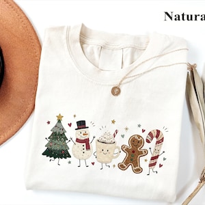 May include: A natural-colored t-shirt featuring a graphic print of a decorated Christmas tree, a snowman with a red scarf, a mug of hot cocoa with whipped cream, a gingerbread man, and a candy cane, all with small arms and legs.