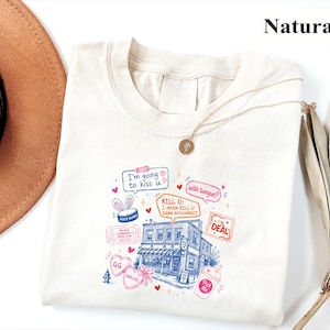 May include: A natural-colored t-shirt featuring a colorful graphic print of a building, speech bubbles with text including "I'm going to kiss u" and "KILL U: I MEAN KILL U DAMN AUTOCORRECT," heart shapes, a ticket, and a drink cup illustration.
