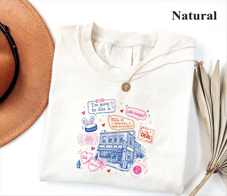 May include: A natural-coloured t-shirt featuring a colourful graphic print of a building, speech bubbles with text including "I'm going to kiss u" and "KILL U: I MEAN KILL U DAMN AUTOCORRECT," heart shapes, a ticket, and a drink cup illustration.