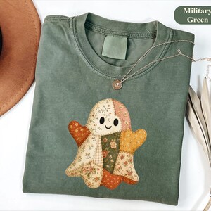 May include: A military green t-shirt featuring a central graphic of a ghost composed of various floral and geometric patchwork patterns in shades of orange, cream, and green. The ghost has two black oval eyes and a small black curved smile.