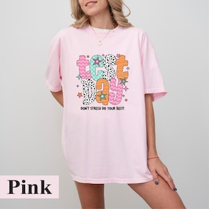 May include: Pink t-shirt with the words "Test Day" in colorful, playful lettering. The design includes stars and the phrase "Don't Stress! Do Your Best!" in a whimsical font. The shirt is a light pink color.