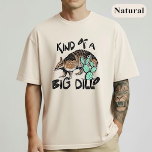 May include: Cream-colored t-shirt featuring a graphic of an armadillo and a cactus with the text "KIND OF A BIG DILLO" in black. The shirt has short sleeves and a crew neck.