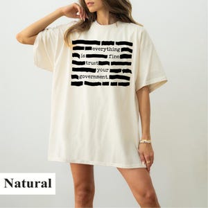 Può includere: T-shirt oversize color naturale con la scritta "everything is fine, trust your government" in nero. La maglietta ha maniche corte e una vestibilità ampia.