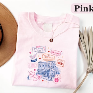 May include: A light pink t-shirt featuring a multi-coloured graphic print of a building, hearts, and text including "I'm going to kiss u", "with tongue?", "KILL U: I MEAN KILL U DAMN AUTICORRECT", "HOCKEY ROMANCE BOOK CLUB", "THE DEAL", "GG", "WELLSY", and "Pick Me".