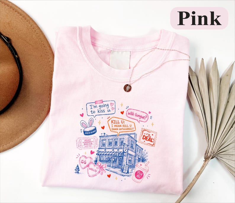 May include: A light pink t-shirt featuring a multi-coloured graphic print of a building, hearts, and text including "I'm going to kiss u", "with tongue?", "KILL U: I MEAN KILL U DAMN AUTICORRECT", "HOCKEY ROMANCE BOOK CLUB", "THE DEAL", "GG", "WELLSY", and "Pick Me".