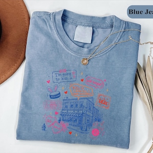 May include: A blue jean coloured t-shirt featuring a graphic print of a building surrounded by various speech bubbles and text including "I'm going to kiss u," "KILL U," and "DEAL." The shirt has a crew neckline and a blank white interior neck label.