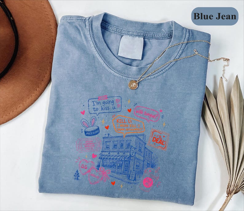 May include: A blue jean coloured t-shirt featuring a graphic print of a building surrounded by various speech bubbles and text including "I'm going to kiss u," "KILL U," and "DEAL." The shirt has a crew neckline and a blank white interior neck label.