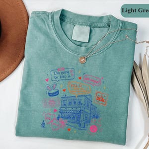 May include: A light green t-shirt featuring a graphic print of a building, hearts, and text including "I'm going to kiss u", "KILL U", and "THE DEAL". The shirt has a crew neckline and a blank label at the collar. The design includes various small icons like a bunny-eared hat, a fire hydrant, and speech bubbles in pink, orange, and blue ink.