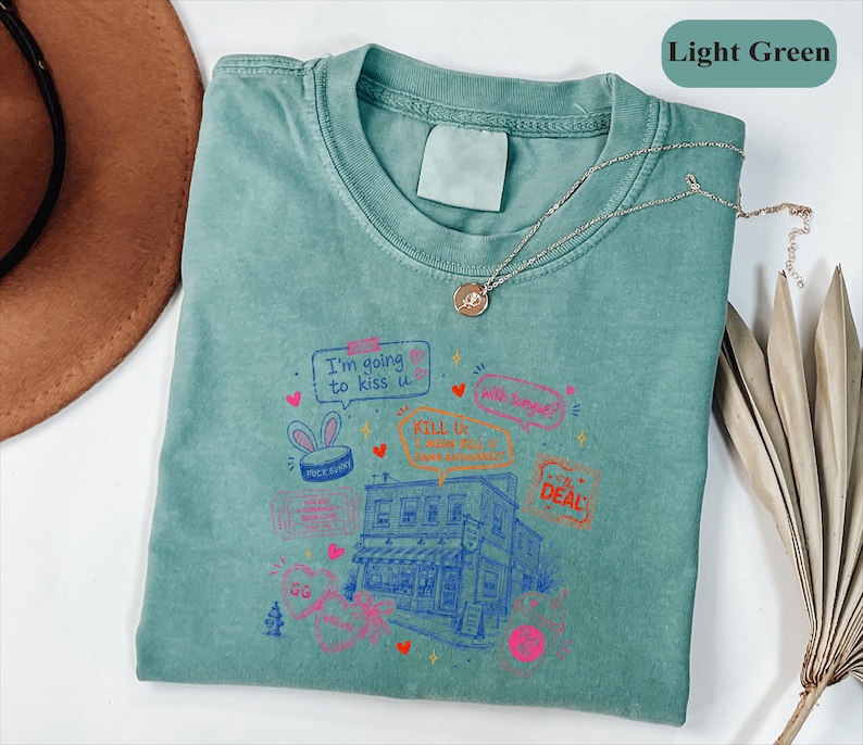 May include: A light green t-shirt featuring a graphic print of a building, hearts, and text including "I'm going to kiss u", "KILL U", and "THE DEAL". The shirt has a crew neckline and a blank label at the collar. The design includes various small icons like a bunny-eared hat, a fire hydrant, and speech bubbles in pink, orange, and blue ink.