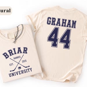 May include: A natural-colored t-shirt featuring a navy blue graphic on the front that reads BRIAR UNIVERSITY HAWKS EST. 2015 with crossed hockey sticks and a puck. The back of the shirt displays the name GRAHAM and the number 44 in navy blue text.