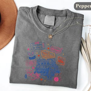 May include: A charcoal grey t-shirt featuring a graphic print of a building with various speech bubbles and text including "I'm going to kiss u" and "KILL U I MEAN KILL U". The design includes small hearts, stars, and a bunny ear icon in pink, blue, and orange.