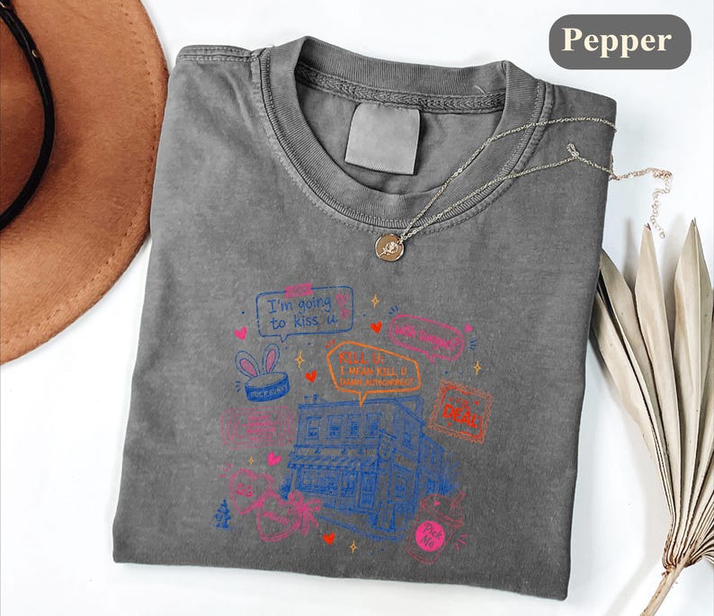 May include: A charcoal grey t-shirt featuring a graphic print of a building with various speech bubbles and text including "I'm going to kiss u" and "KILL U I MEAN KILL U". The design includes small hearts, stars, and a bunny ear icon in pink, blue, and orange.