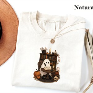 May include: A natural-colored t-shirt featuring a graphic of a ghost sitting at a small round table, writing in a book with a quill. The scene includes a bookshelf, pumpkins, a black cat, a skull, candles, and autumn foliage.