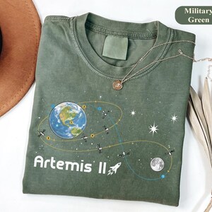 Könnte beinhalten: Militärgrünes T-Shirt mit einer Grafik der Erde, Satelliten, des Mondes und Sterne. Der Text "Artemis II" und eine Rakete sind darunter gedruckt. Eine goldene Halskette liegt über dem Shirt.