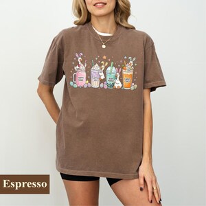 May include: Espresso-colored t-shirt featuring a colorful graphic of Easter-themed drinks. The design includes bunny characters in various beverages with text like "Hoppy Shake" and "Carrot Craze". The shirt is a casual, short-sleeved style.