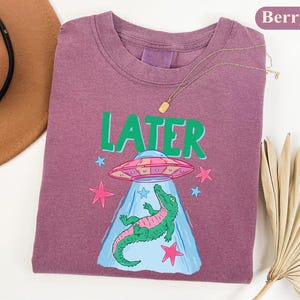 May include: Berry-coloured t-shirt with a crew neck. The shirt features a graphic of a green alligator being abducted by a UFO, with the word "LATER" in green. Pink stars and a blue beam complete the design.