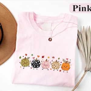 Pode incluir: Uma t-shirt rosa claro com uma estampa gráfica de cinco abóboras de desenho animado com vários padrões, incluindo bolinhas, estampa de leopardo e cores sólidas, acentuada por pequenas folhas de outono, corações e um ícone de cogumelo.