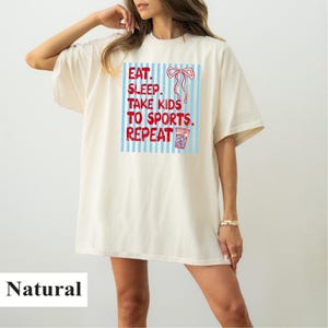 Op de afbeelding: Crèmekleurig T-shirt met de tekst "EAT. SLEEP. TAKE KIDS TO SPORTS. REPEAT" in rood en blauw. De afbeelding bevat een strik en een drankje. Het shirt heeft een losse pasvorm.