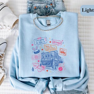 May include: A light blue crewneck sweatshirt featuring a graphic print of a building surrounded by various doodles, including speech bubbles with the text "I'm going to kiss u", "with tongue?", and "KILL U: I MEAN KILL U DAMN AUTOCORRECT".