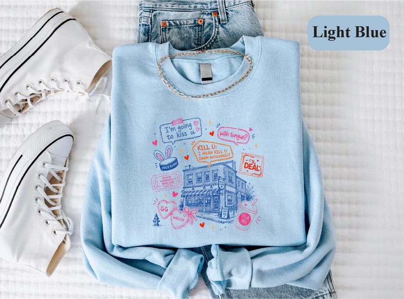 May include: A light blue crewneck sweatshirt featuring a graphic print of a building surrounded by various doodles, including speech bubbles with the text "I'm going to kiss u", "with tongue?", and "KILL U: I MEAN KILL U DAMN AUTOCORRECT".