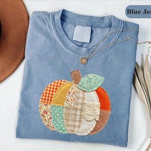 May include: A blue jean colored t-shirt featuring a central pumpkin applique made from various fabric patches including orange gingham, floral print, green polka dots, plaid, and lace, with a gold tone necklace resting on the collar.