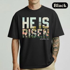 Puede incluir: Camiseta negra con las palabras "HE IS RISEN" en un diseño floral. El texto "Matthew 28:6" está debajo del texto principal. La palabra "Black" está en un cuadro blanco en la esquina superior derecha.