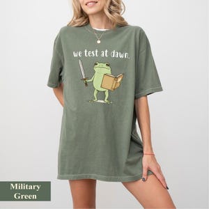 May include: Military green t-shirt with the text "we test at dawn." The shirt features a cartoon frog holding a sword and a book. The frog is light green, the sword is silver, and the book is tan.