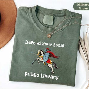 May include: Military green t-shirt featuring the text "Defend Your Local Public Library" and a graphic of a knight on horseback. The shirt has a crew neck and short sleeves. A gold necklace with a pendant is draped across the shirt.